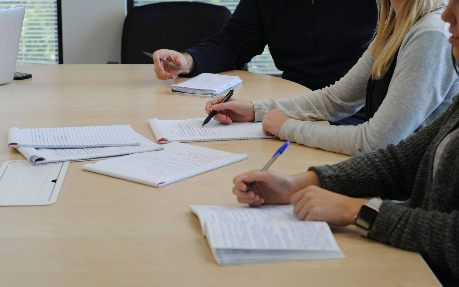 Three people at a meeting table writing in notebooks, with printed pages spread between them